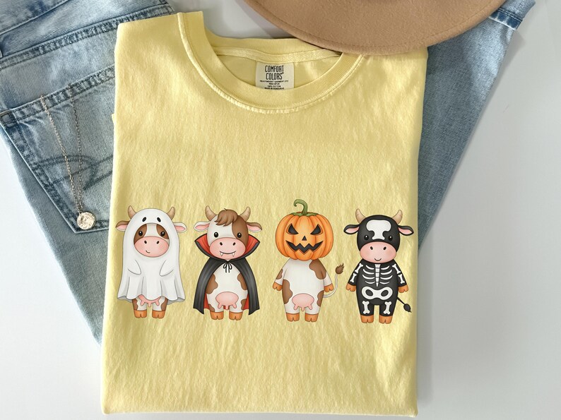 May include: Yellow Comfort Colors t-shirt featuring four cartoon cows in Halloween costumes: a ghost, a vampire, a jack-o'-lantern, and a skeleton. The shirt is laid flat on a light surface with a pair of blue jeans and a necklace.