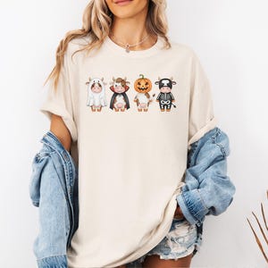 May include: Cream-colored t-shirt featuring four cartoon cows dressed in Halloween costumes: a ghost, a vampire, a jack-o'-lantern, and a skeleton. The cows are in a row, each with unique details. The shirt is a casual, unisex style.