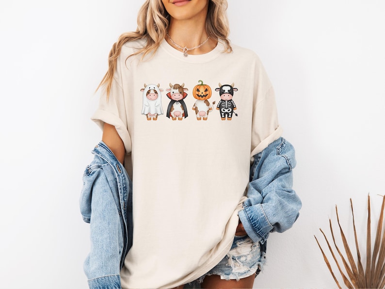 May include: Cream-colored t-shirt featuring four cartoon cows dressed in Halloween costumes: a ghost, a vampire, a jack-o'-lantern, and a skeleton. The cows are in a row, each with unique details. The shirt is a casual, unisex style.