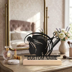 Peut inclure: Une silhouette en métal noir représentant une coiffure, des ciseaux et un peigne, fixée sur une base rectangulaire en bois clair. La base porte l'inscription CUSTOM NAME en lettres capitales noires, centrée sous le motif.