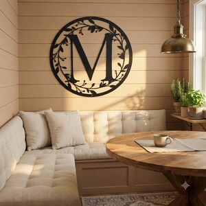 May include: A circular black metal wall sign featuring a large capital letter M centred within a decorative frame of thin branches and leaves. The sign is mounted on a horizontal wood-panelled wall above a tufted beige bench seat.