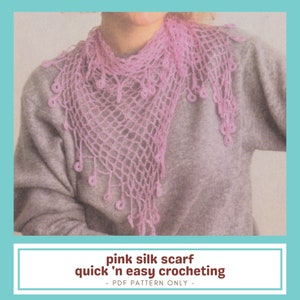 May include: A light pink, crocheted silk scarf with a delicate net-like design and dangling circular accents. The scarf is triangular in shape and is displayed on a person wearing a gray sweater. The text on the image reads: "pink silk scarf quick 'n easy crocheting".