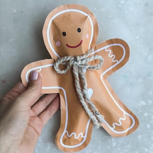 May include: A gingerbread man-shaped package with a smiling face, pink cheeks, and a white outline. The package is tied with a natural twine bow. The overall color is a light brown.