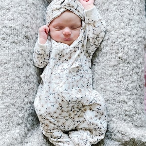 Pode incluir: Um bebê vestindo um macacão branco com estampa floral e uma faixa de turbante branca. O bebê está deitado em um cobertor cinza felpudo.