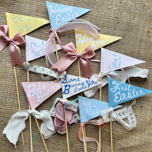 May include: A set of ten pastel-coloured pennant flags with Easter-themed designs and text. The flags are decorated with ribbons and lace. The text on the flags includes "Happy Easter", "Hip Hop", "Baby Bunny", "Some Bunny Loves You", and "First Easter".