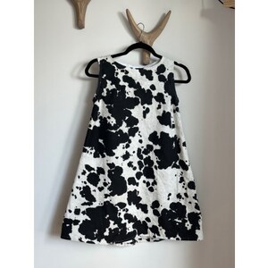 May include: Sleeveless dress with a cow print design. The dress features a white background with irregular black spots. It hangs on a black hanger against a white wall. The dress is a casual garment.