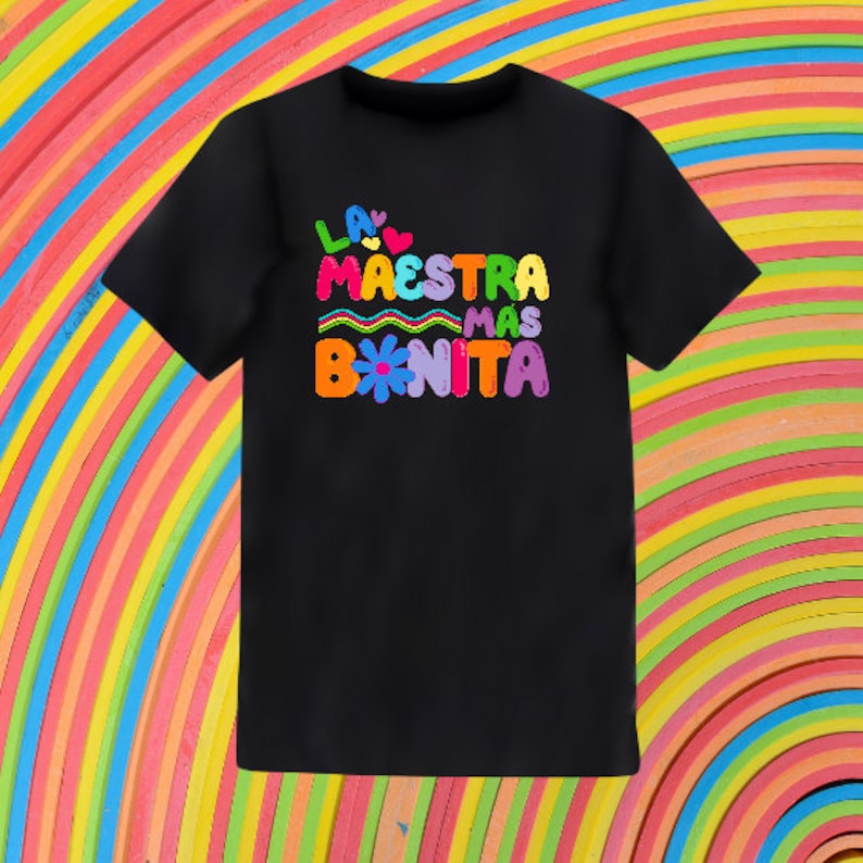 Puede incluir: Camiseta negra con un gr&aacute;fico colorido que dice "La Maestra Mas Bonita" con una flor y corazones.