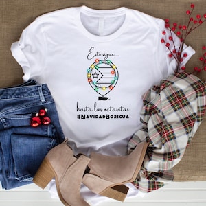 Op de afbeelding: Wit T-shirt met een zwarte afbeelding van een Puerto Ricaanse vlag met kerstverlichting eromheen. De tekst "Esto sigue... hasta las octavitas #NAVIDADBORICUA" staat onder de afbeelding.