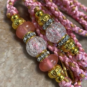 May include: A pink and gold braided cord with clear and pink glass beads. The beads are accented with gold and rhinestones.