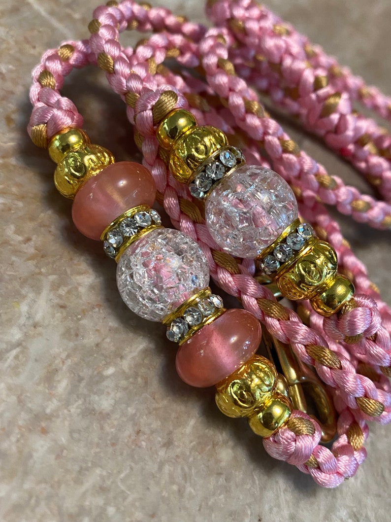May include: A pink and gold braided cord with clear and pink glass beads. The beads are accented with gold and rhinestones.