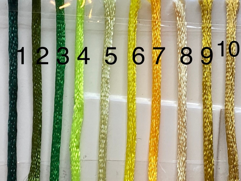 May include: Ten strands of embroidery floss in shades of green and yellow. The floss is numbered 1 through 10.