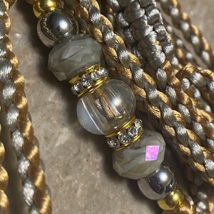 May include: A close-up of a gold and silver braided cord with crystal beads and gold accents. The cord is made of two strands of braided cord, one gold and one silver. The beads are clear and have a faceted design. The gold accents are small and round.