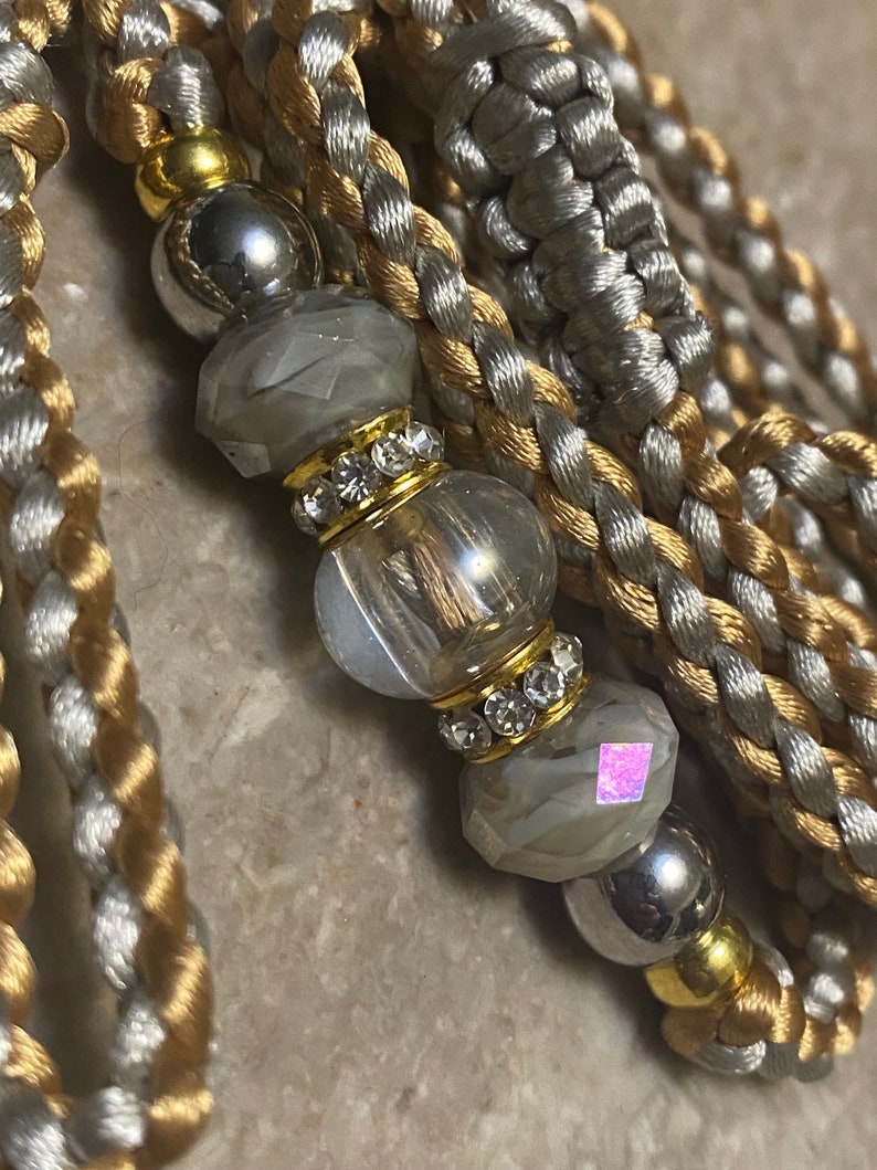 May include: A close-up of a gold and silver braided cord with crystal beads and gold accents. The cord is made of two strands of braided cord, one gold and one silver. The beads are clear and have a faceted design. The gold accents are small and round.