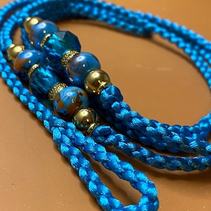 May include: A blue braided cord with gold and blue beads. The cord is twisted and has a tassel at the end.