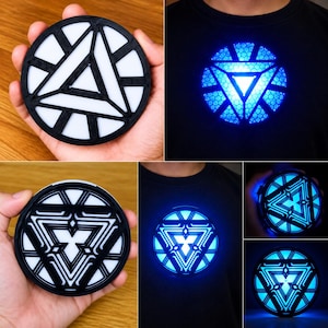 May include: A circular arc reactor prop featuring a black frame with white geometric inserts. The device emits a bright blue light when activated and is shown attached to a black t-shirt, displaying intricate triangular and linear patterns.