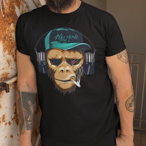 Könnte beinhalten: Schwarzes T-Shirt mit einem Affen-Grafik, der Kopfhörer und eine Kappe trägt, mit dem Text "Dirty Music". Der Affe raucht eine Zigarette. Das Design verwendet Braun-, Türkis- und Schwarztöne.