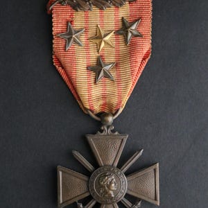 Puede incluir: Medalla militar de bronce con cinta a rayas rojas, amarillas y blancas. La medalla presenta un diseño de cruz con espadas y un medallón central con un perfil. Encima de la cinta hay cuatro estrellas y una hoja de bronce.