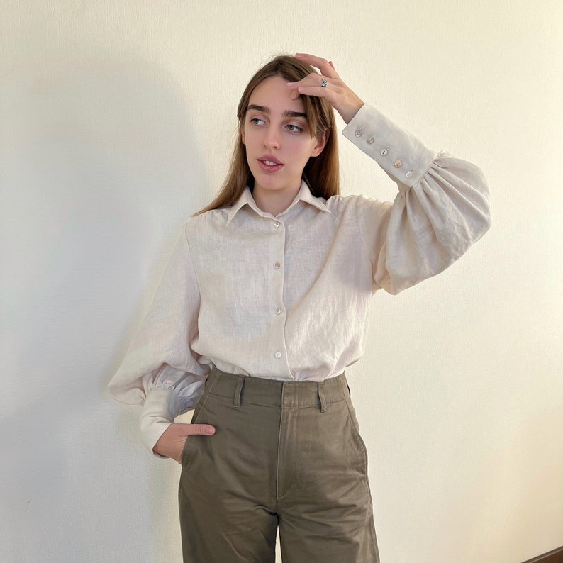 May include: A woman wearing a light beige linen shirt with puffed sleeves and a button-down collar. She is also wearing olive green high-waisted pants.