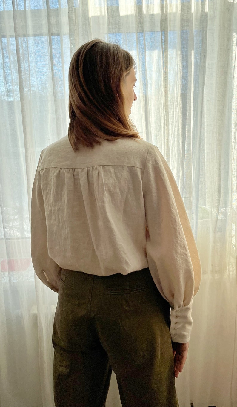 May include: A woman wearing a white linen blouse with long sleeves and a gathered back detail. She is also wearing dark green pants.