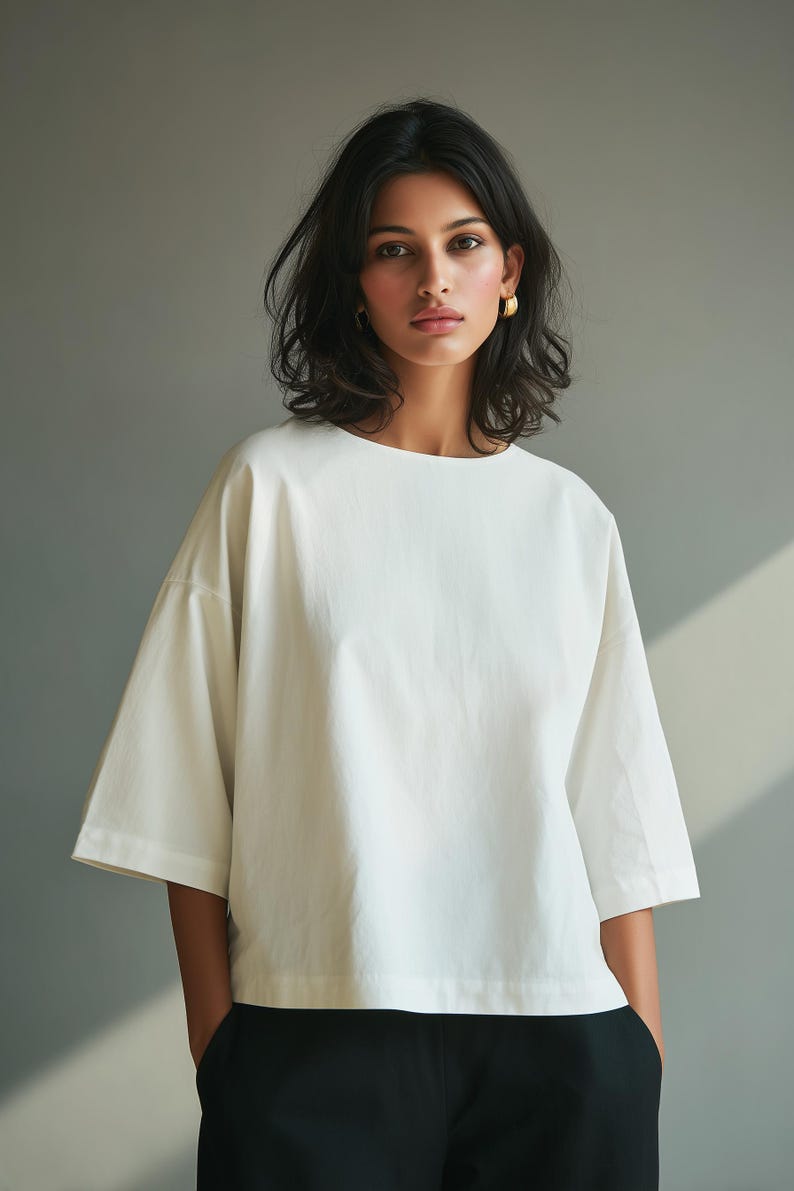 May include: A woman wearing a white, short-sleeved top with a boat neck and black pants. The top has a loose fit. The woman has dark hair and is wearing gold earrings. The background is a solid gray.