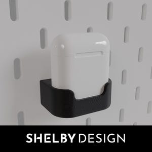Peut inclure: Un étui de charge sans fil blanc pour écouteurs est monté sur un support mural imprimé en 3D noir. Le support est conçu pour s'adapter à un mur en panneau perforé.