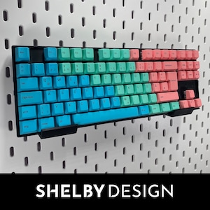 Könnte beinhalten: Eine Tastatur mit schwarzem Rahmen und blauen und rosa Tasten. Die Tasten haben weiße Beschriftung. Die Tastatur ist an einer Lochwand montiert.