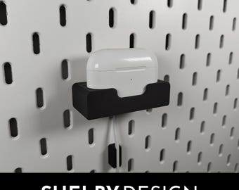 May include: A white wireless charging case for earbuds is mounted on a black wall mount. The mount is attached to a pegboard wall. The text "SHELBY DESIGN" is visible at the bottom of the image.