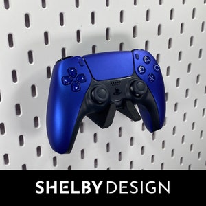 May include: A blue PlayStation 5 DualSense controller mounted on a wall with a pegboard background. The controller has a black faceplate and black buttons.