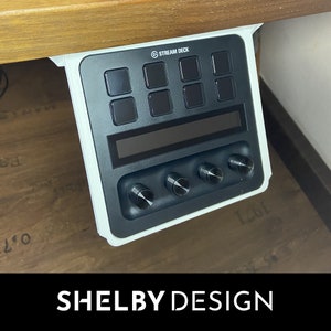May include: A black Stream Deck control panel featuring eight square buttons, a display screen, and four rotary knobs. It's mounted beneath a wooden surface with a white bracket. The text "STREAM DECK" is at the top. The text "SHELBY DESIGN" is at the bottom.