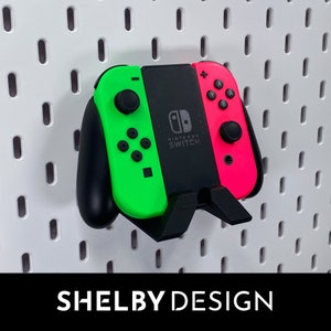 Peut inclure: Une manette Nintendo Switch verte et rose avec un support noir. La manette est fixée à un mur en panneau perforé blanc. Le texte "NINTENDO SWITCH" est imprimé sur la manette.