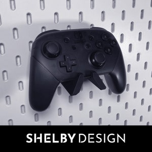 Puede incluir: Un mando negro de Nintendo Switch con un soporte de pared negro. El mando tiene el logotipo de Nintendo Switch y botones etiquetados como X, Y, A y B. El soporte de pared está diseñado para sujetar el mando de forma segura.