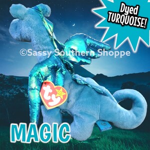May include: A plush dragon toy with light blue fur and metallic turquoise wings. It features a heart-shaped red Ty tag attached to the chest and a blue tag on the tail. The text "Dyed TURQUOISE!" and "MAGIC" are visible, along with "Sassy Southern Shoppe".
