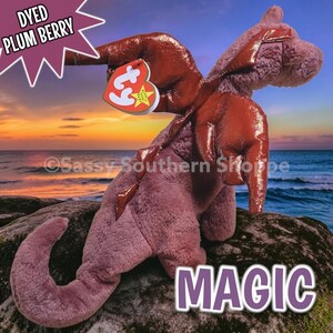 May include: A plush dragon toy in a muted plum berry colour with shiny, metallic-textured wings. The stuffed animal features a heart-shaped tag with the text ty attached to the wing. The word MAGIC is printed in large white letters at the bottom.