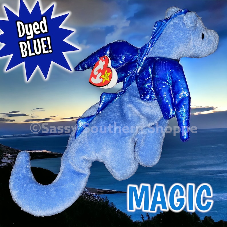 May include: A light blue plush dragon toy with shiny dark blue wings and a heart-shaped Ty tag attached. The toy features the text Dyed BLUE! in a starburst graphic and the word MAGIC in large blue letters against a coastal sunset background.