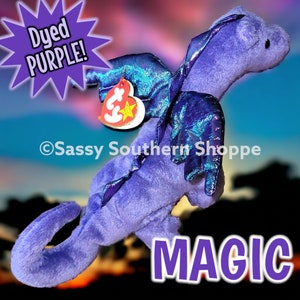 May include: A purple plush dragon toy with iridescent blue wings and a heart-shaped Ty tag attached. The image includes the text Dyed PURPLE!, Sassy Southern Shoppe, and MAGIC. The dragon has a soft texture, a long tail, and small dark eyes.