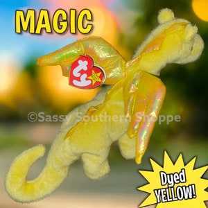 May include: A yellow plush dragon toy with iridescent wings and a heart-shaped red Ty tag attached. The word MAGIC is printed in yellow at the top, and a yellow starburst graphic at the bottom contains the text Dyed YELLOW!