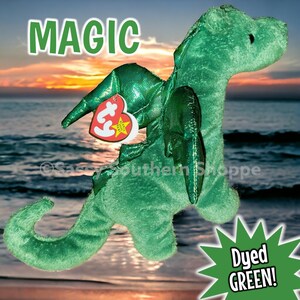May include: A green plush dragon toy with a textured surface, featuring a heart-shaped red and white Ty Beanie Baby tag attached to its wing. The word MAGIC is printed in green at the top, and a green starburst graphic says Dyed GREEN!