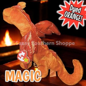 May include: An orange plush dragon toy with a textured fabric body and shiny, metallic-finish wings. A heart-shaped red Ty tag is attached to the chest. The image includes the text "Dyed ORANGE!" in a starburst and the word "MAGIC" at the bottom.