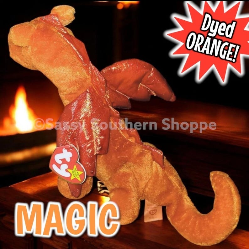 May include: An orange plush dragon toy with a textured fabric body and shiny, metallic-finish wings. A heart-shaped red Ty tag is attached to the chest. The image includes the text "Dyed ORANGE!" in a starburst and the word "MAGIC" at the bottom.