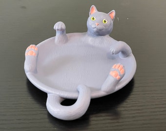 May include: A grey ceramic cat dish featuring a cat figurine lying on its back with its paws and tail sticking out. The cat has yellow eyes and pink paw pads.