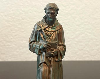 May include: A bronze statue of a robed figure holding a book and a cross. The statue has a weathered appearance with green and brown tones. The figure's face is detailed, and the statue is set against a neutral background.