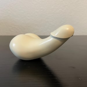 Peut inclure: Une sculpture en forme de phallus de couleur crème, avec une base arrondie et une tige incurvée. La sculpture est lisse et semble être faite de céramique ou d'un matériau similaire. L'objet est posé sur une surface sombre, avec un mur clair en arrière-plan.
