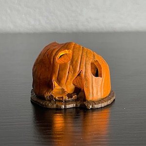May include: A small, orange, ceramic pumpkin with a carved face and a lit candle inside. The pumpkin is sitting on a brown base.