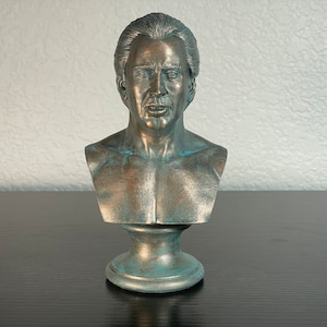 Pode incluir: Um busto de bronze de um homem com acabamento envelhecido. O busto está em um pedestal e tem uma expressão realista.