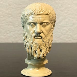 May include: A cream-colored bust of a bearded man with a stern expression. The sculpture features detailed facial features, including a prominent nose and a full beard. The bust is mounted on a circular base.