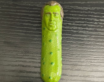Peut inclure: Un cornichon vert avec un visage humain, ressemblant à Nicolas Cage, est présenté sur un fond en bois noir.