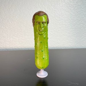 May include: A green pickle with a human face on top. The pickle is standing on a white base.