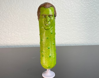 Peut inclure: Un cornichon vert avec un visage humain sur le dessus. Le cornichon est posé sur une base blanche.
