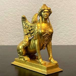 Puede incluir: Estatua de esfinge dorada con cabeza de mujer, alas y cuerpo de león. La estatua está sobre una base rectangular y presenta detalles intrincados, incluyendo un tocado y elementos decorativos en el pecho. Un artículo decorativo.