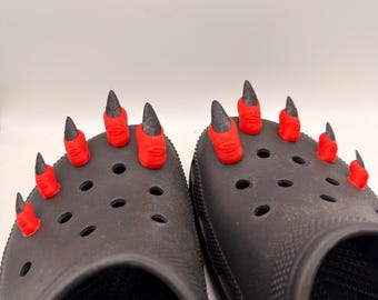 May include: A pair of black clogs adorned with red and black claw charms. The charms are designed to resemble devil claws, with red nail beds and black pointed nails. The clogs have multiple ventilation holes.